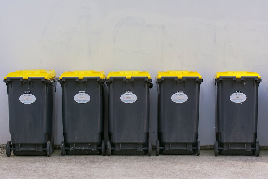 Five large wheeled rubbish or waste collection bins lined up against a plain, light-colored wall, with the bins evenly spaced on a concrete ground. The bins are made of sturdy black plastic and each features a bright yellow lid, which appears to be made of hard, durable material with a smooth, slightly reflective surface. Small white labels with black text and barcodes are affixed to the front of each bin, indicating their identification numbers. The bins are positioned in a neat row, with their wheels aligned, and are set in an outdoor area, possibly a driveway or service yard. The ambient lighting is diffuse, suggesting an overcast day, and the environment appears clean and orderly. This scene reflects aspects of on-site waste management and potential private waste disposal options, as provided by companies like Rubbish Clearance Uxbridge, offering an alternative to traditional council-collected rubbish services.