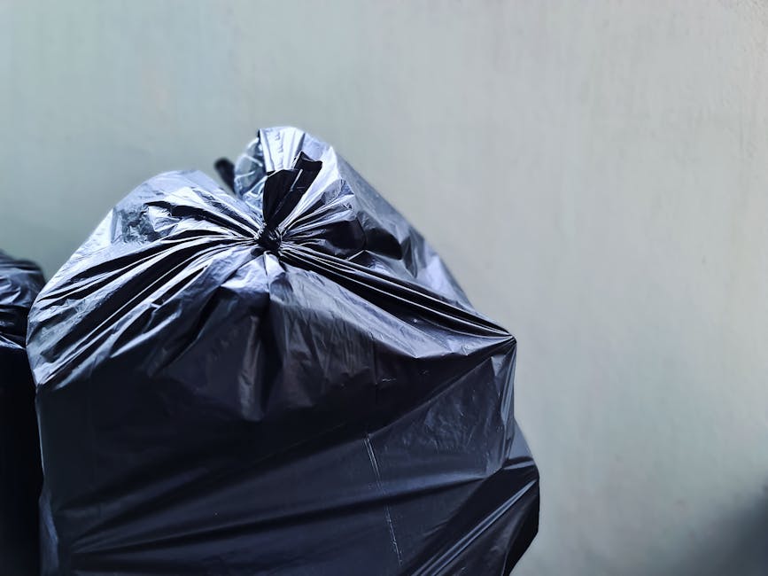 A close-up view of multiple black plastic rubbish bags tightly tied at the top, stacked together in a disposal area or on a pavement, with a plain light-colored wall in the background. The bags appear to be made of glossy plastic material with visible creases and folds, indicating they are filled with waste. The bags are positioned on the left side of the image, with some partially overlapping, and are illuminated evenly, highlighting the reflective surface of the plastic. This scene reflects typical private waste disposal or on-site clearance activities often handled by independent rubbish removal services like Rubbish Clearance Uxbridge. The environment suggests an outdoor location near a building or behind a property, emphasizing the context of waste collection outside of conventional municipal rubbish services.