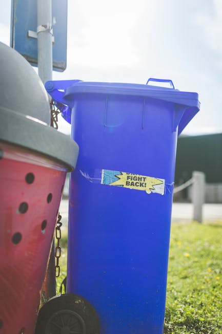 Five large wheeled rubbish or waste collection bins lined up against a plain, light-colored wall, with the bins evenly spaced on a concrete ground. The bins are made of sturdy black plastic and each features a bright yellow lid, which appears to be made of hard, durable material with a smooth, slightly reflective surface. Small white labels with black text and barcodes are affixed to the front of each bin, indicating their identification numbers. The bins are positioned in a neat row, with their wheels aligned, and are set in an outdoor area, possibly a driveway or service yard. The ambient lighting is diffuse, suggesting an overcast day, and the environment appears clean and orderly. This scene reflects aspects of on-site waste management and potential private waste disposal options, as provided by companies like Rubbish Clearance Uxbridge, offering an alternative to traditional council-collected rubbish services.