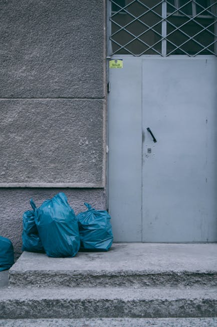 The image shows three bright blue rubbish bags placed on a concrete step in front of a building's exterior wall, which has a rough, textured grey surface. To the right of the bags, there is a grey metal door fitted with a black handle, situated on a concrete platform. The door has a small lock below the handle and features a grid-like window at the top, composed of metal bars forming a diamond pattern, with glass panes behind. A small yellow warning or information sticker is affixed near the top of the door frame. The environment appears urban, with natural daylight illuminating the scene, suggesting the bags are temporarily stored outside, possibly awaiting collection as part of private waste disposal services by Rubbish Clearance Uxbridge. The setting emphasizes an on-site rubbish removal context, aligning with alternative waste handling methods that bypass local authority collections.