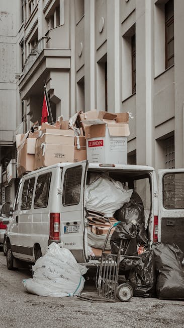 A white commercial van parked on a city street with its rear doors open, revealing a large load of mixed waste materials including black and white plastic rubbish bags, cardboard boxes, and wooden pallets. The van is positioned next to a tall, multi-storey building with a beige facade, featuring several large windows and architectural details such as moldings and protruding ledges. Cardboard boxes, some with printed labels, are stacked on top of the van, indicating the collection of household or commercial waste for private disposal. In front of the van, a small trolley cart loaded with black rubbish bags and loose debris stands on the pavement, surrounded by scattered waste. The scene appears as an outdoor rubbish clearance operation, typical of a professional waste removal service handling bulky or mixed rubbish outside urban premises, aligning with the context of independent waste collection or on-site clearance. The natural ambient light highlights the textures of the waste materials and the building's detailed exterior, emphasizing a clean and professional waste management approach without any promotional language.