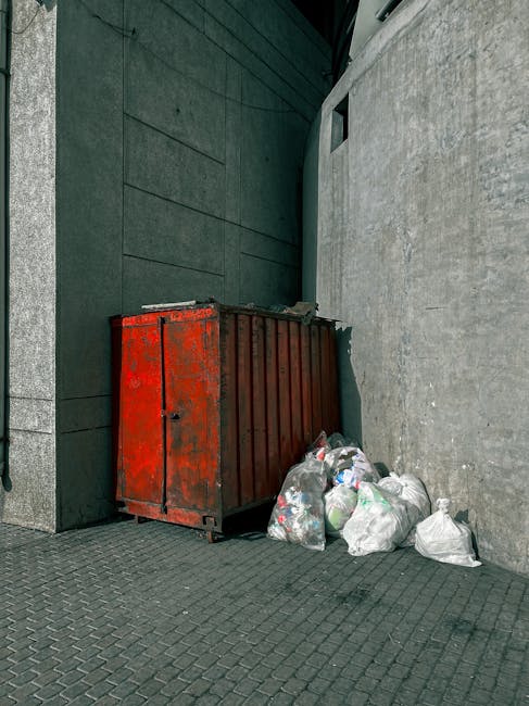 A red metal skip container with a weathered and slightly rusted surface, positioned against the base of a modern dark green wall on the left side and a light grey concrete wall on the right. In front of the skip, there are several clear plastic garbage bags filled with mixed waste, including visible plastic bottles and paper. The ground is paved with dark grey interlocking bricks, and the scene appears to be located on a paved alleyway or backstreet area. The surrounding environment suggests an external urban setting, possibly near a commercial or residential building, with minimal natural lighting, creating a neutral, utilitarian atmosphere. This scene by Rubbish Clearance Uxbridge illustrates typical waste accumulation outside a structure needing collection or clearance, aligning with independent disposal practices outside council-controlled rubbish collection points, reflecting private waste handling options in the context of rubbish removal services.