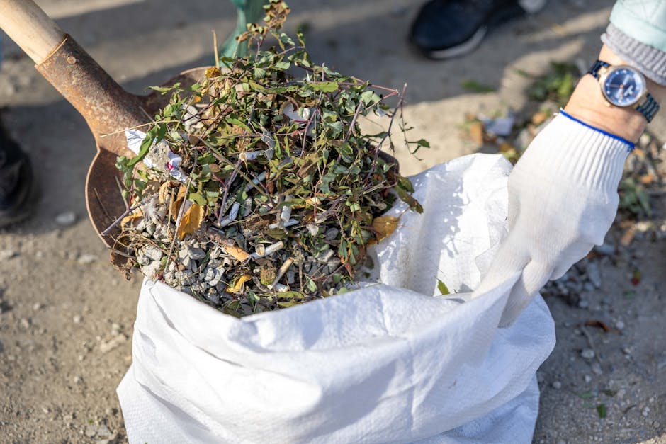 A close-up view of a white plastic sack filled with a mixture of garden and outdoor waste, including small leafy branches, twigs, and scattered gravel. The sack is held open by a person wearing a grey and white striped glove on their right hand, which is visible gripping the edge of the bag. A small section of a metal shovel with a rusted surface is positioned on the left side, resting against the waste, with the handle partly out of the frame. The background shows a patch of pavement or dirt ground with some greenery and additional small debris scattered nearby. The scene appears to be outdoors, likely during a rubbish removal or clearance activity, which aligns with services offered by Rubbish Clearance Uxbridge, focusing on private waste disposal and environmentally responsible rubbish collection. The natural lighting highlights the textures of the gravel, branches, and the fabric of the sack, emphasizing the details of the waste material intended for disposal or recycling.