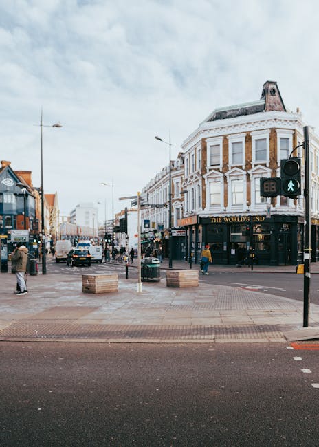 A busy urban street scene featuring a prominent Victorian-style corner building with white and beige brickwork, large rectangular windows, and a sloped roof with a chimney. The building houses a pub named 'The World's End,' situated at the intersection of pavements and roads. In the foreground, there are concrete curb extensions with embedded wooden planters, some with small plants, and pedestrian crossings marked on the asphalt, with traffic signals showing a green pedestrian icon. The street is populated with pedestrians walking, some waiting at crosswalks, and vehicles such as cars and vans driving along the road. Overhead, a partly cloudy sky is visible, with streetlights and lampposts lining the pavement. The scene captures a typical city centre environment, where local businesses and public transport co-exist, and the setting aligns with contexts where independent or alternative waste handling might occur, as managed by local rubbish clearance services like Rubbish Clearance Uxbridge.